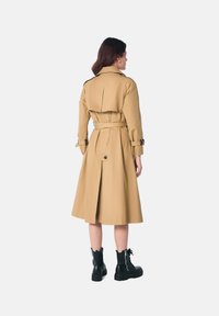 Trench-coat beige avec une taille ceinturée, style double-breasted et une fente à l'arrière. Porté avec des bottes de combat noires. Tissu texturé.