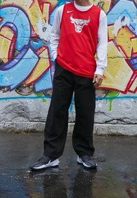 Jersey sin mangas rojo con gráfico de toro, mangas largas blancas, pantalones anchos negros y zapatillas grises frente a un colorido fondo de graffiti.