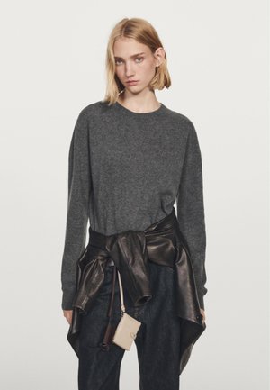 Mørkeblå sweater med lange ærmer, parret med et bronze læderbælte om taljen. Blå denimbukser og en lille beige pung som accessory.