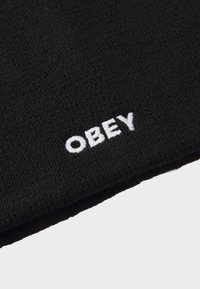 Črna pletena kapa z teksturirano tkanino. Ima bel vtičeni logotip "OBEY" na robu. Preprost, neokrašen oblik.
