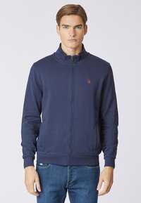 Felpa zip-up blu navy realizzata in tessuto liscio, con colletto alto, polsini a costine e un piccolo logo rosso sul petto.