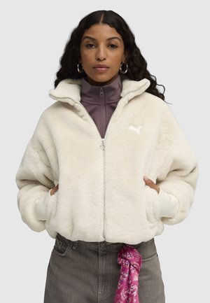Weißer, flauschiger Reißverschlussjacke mit hohem Kragen, ausgestattet mit einer weichen Textur und gerippten Bündchen, über einem lila hochkragen Oberteil getragen.