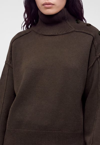 Femme portant un pull à col roulé côtelé marron foncé avec des détails de boutons sur l'épaule et des manches longues.