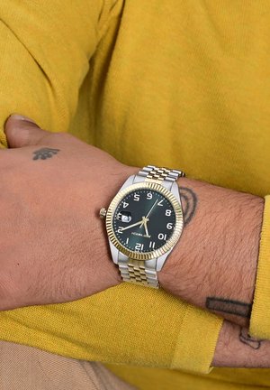 Gold- und silberfarbene Uhr mit schwarzem Zifferblatt, großen weißen Zahlen, Datumsfenster und zweifarbigem Metallarmband. Getragen an einem Männerhandgelenk.