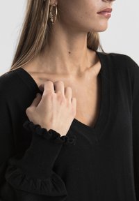 Haut en maille noire avec encolure en V, doté de poignets noirs à volants. La main du mannequin repose sur le tissu près de l'encolure. Texture lisse.