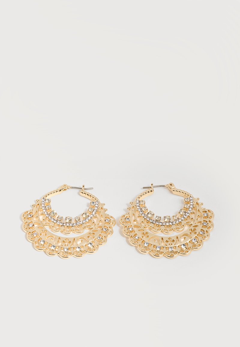 Paire de boucles d’oreilles créoles dorées avec des motifs complexes en forme de dentelle et des strass clairs incrustés sur fond blanc.