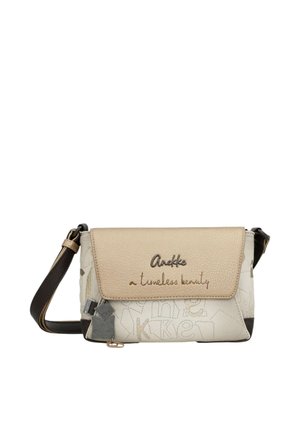Piccola borsa a tracolla beige e bianca con tracolla nera, chiusura a patta con testo ricamato "Anekke a timeless beauty" e charm decorativi.