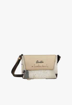 Piccola borsa a tracolla beige e bianca con tracolla nera, chiusura a patta con testo ricamato "Anekke a timeless beauty" e charm decorativi.