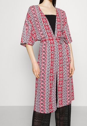 Osoba ubrana w czerwoną, białą i czarną sukienkę typu wrap z półdługimi rękawami i przewiązanym w talii paskiem, na czarnych koronkowych spodniach.