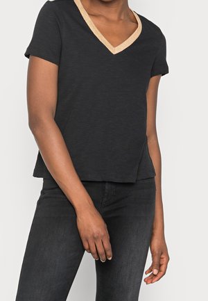 Kobieta ubrana w czarny T-shirt z krótkim rękawem i dekoltem w serek z metaliczną, złotą lamówką, w połączeniu z czarnymi dopasowanymi jeansami, stojąca z rozluźnionymi rękami.