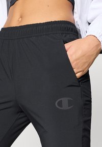 Juodi sportiniai kelnių su tampria juosmeniu, šoninėmis kišenėmis ir lygiu paviršiumi. Diskretiškai logotipas yra atspausdintas ant apatinės kojos.