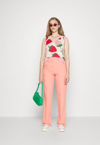 Colete de malha com padrão de morangos, combinado com calças rosa claras. Os acessórios incluem uma mala verde e óculos de sol redondos rosa.