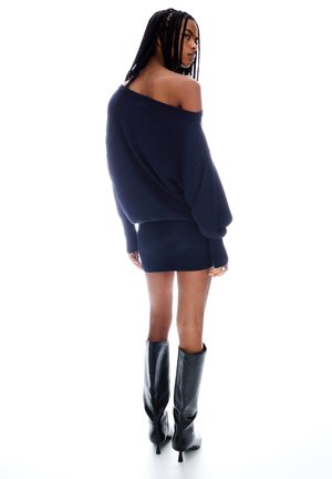 Navy oversized off-shoulder trui, geribbelde rok en zwart schouderlaarzen met spitse neus. Zachte stof, relaxte silhouet en textuurcontrast.