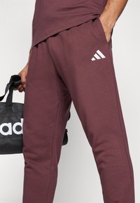 Bordo sporta bikses ar mīkstu tekstūru, ar baltu adidas logotipu un kabatām. Kopā ar melnu somu, kurā redzams zīmola nosaukums.