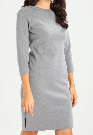 Kobieta w dopasowanej, szarej, dzianinowej sukience z długim rękawem sięgającej do kolan, z małym rozcięciem po boku, na białym tle.