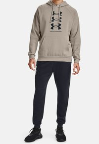Uomo che indossa un hoodie beige di Under Armour con logo e pantaloni sportivi neri con logo sulla coscia, abbinati a scarpe da ginnastica nere.
