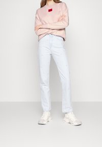 Felpa rosa chiaro con una targhetta rettangolare rossa, abbinata a jeans azzurri a gamba dritta con delicati motivi ricamati. Scarpe da ginnastica bianche.