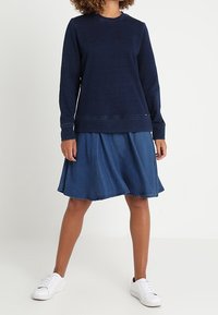 Marineblaues Sweatshirt mit langen Ärmeln, rundem Halsausschnitt und gerippten Bündchen, kombiniert mit einem Jeansrock in ausgestellter Form und rohem Saum. Weiße Sneakers.