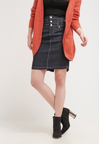 Falda de mezclilla con un diseño ajustado, color azul oscuro, cierre frontal con botones y acentos metálicos, y costuras claras. Combinada con botas negras de tobillo.