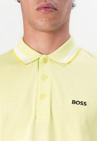 Žlté polo tričko vyrobené z textúrovanej látky, s trojbuttonovou légou a kontrastným bielym a žltým pruhovaným golierom. Čierne logo "BOSS".