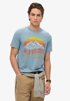 T-shirt en coton bleu chiné avec un graphique de montagnes et d'un coucher de soleil, associé à un pantalon khaki et un pull gris noué à la taille.