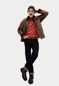 Chaqueta marrón, suéter de punto naranja, jeans oscuros, botas de senderismo marrón y negro. El modelo lleva un gorro de punto azul, de pie con una mano en la cabeza.
