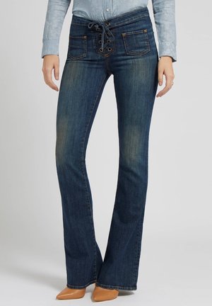 Jean flare bleu foncé avec fermeture à lacets à l'avant et poches, porté avec une chemise bleu clair et des talons pointus beige.