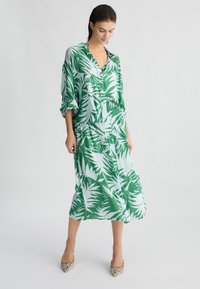 Abito in seta verde e bianco con motivo di foglie tropicali, con colletto e bottoni. Presenta una vestibilità ampia e un dettaglio in vita legato.