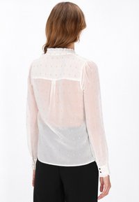 Blusa bianca trasparente con piccoli dettagli in oro e argento, colletto arricciato, polsini con bottoni e vestibilità comoda. Vista posteriore mostrata.