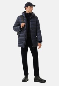 Navy puffer jacket with hood, zip front, and side pockets. Paired with black trousers and ankle boots. Contains a black backpack and cap.
