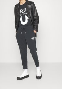 Veste de motard en cuir noir sur un t-shirt noir avec des graphiques blancs, associée à un pantalon de jogging sombre arborant un logo blanc, et des baskets blanches avec des semelles noires.