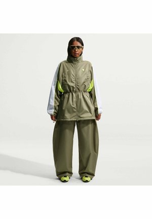 Personen står iført en stor olivengrøn og hvid træningsdragt med neon-gule accenter samt matchende neon-gule sneakers og solbriller.