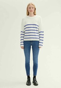 Λευκό πλεκτό πουλόβερ με μπλε οριζόντιες ρίγες, στρογγυλή λαιμόκοψη, μακριά μανίκια. Συνδυασμένο με στενά μπλε τζιν και μαύρα παπούτσια.