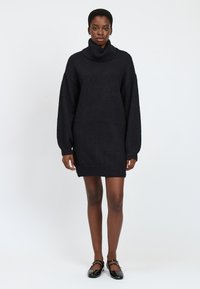Schwarzes, oversized Turtleneck-Kleid aus strukturiertem Strickstoff, mit langen Ärmeln, gerippten Bündchen und Saum, kombiniert mit schwarzen Ballerinas.