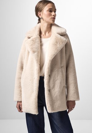 Manteau en fausse fourrure couleur crème avec un large col, coupe décontractée et boutons sur le devant. Associé à un pull léger et un pantalon en denim foncé.