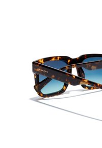 Schildpatt-Sonnenbrille mit blauen Verlaufsgläsern. Dicke Rahmen mit hexagonaler Form, die den Markennamen "Hawkers" an der Seite tragen.