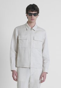 Giacca zip-up color beige chiaro realizzata in lino, con due tasche sul petto, colletto classico e maniche lunghe. Indossata con pantaloni abbinati.