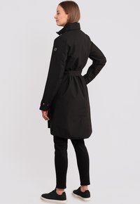 Cappotto trench nero realizzato in un tessuto liscio, con vita cinturata, colletto alto e polsini abbottonati, abbinato a leggings neri e sneakers.