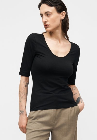 Γυναίκα με τατουάζ φοράει μαύρο τοπ με μισά μανίκια και στρογγυλή λαιμόκοψη και μπεζ παντελόνια, ποζάρει με το ένα χέρι στην τσέπη, κοιτάζοντας προς το πλάι.