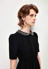 Schwarzes Kleid mit kurzen Ärmeln, das einen mit Perlen bestickten Kragen im Punktemuster aufweist. Glatte Textur, taillierter Oberkörper und schwingender Rock.