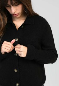 Gilet en laine noir avec un col, des poches avant et de grands boutons. Le tissu semble doux et texturé.