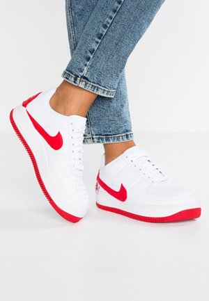Personne portant un jean bleu et des baskets blanches avec des logos Nike rouges et des semelles rouges, debout sur un sol blanc.
