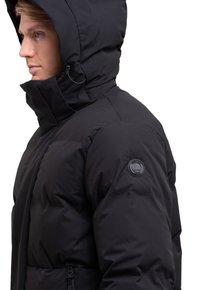 Chaqueta acolchada negra con un cuello alto y capucha. Presenta una cremallera lateral, tejido texturizado y un parche de logo circular en la manga izquierda.