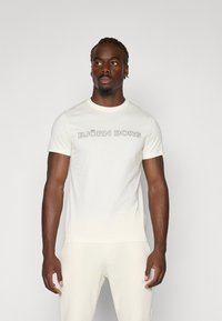 Cremefarbiges T-Shirt mit kurzen Ärmeln, hergestellt aus weichem Stoff, mit dem Text "BJÖRN BORG" in kräftiger schwarzer Serifenschrift auf der Brust.
