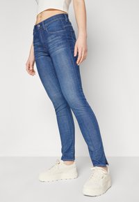 Tynne, blå denimjeans med midjehøyde, subtile sømdetaljer og sidelommer ved kanten. Kombinert med lyse fottøy.
