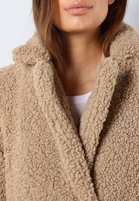 Cappotto teddy beige, testurizzato, con collo a scialle, caratterizzato da un esterno morbido e peloso e pannelli anteriori sovrapposti.