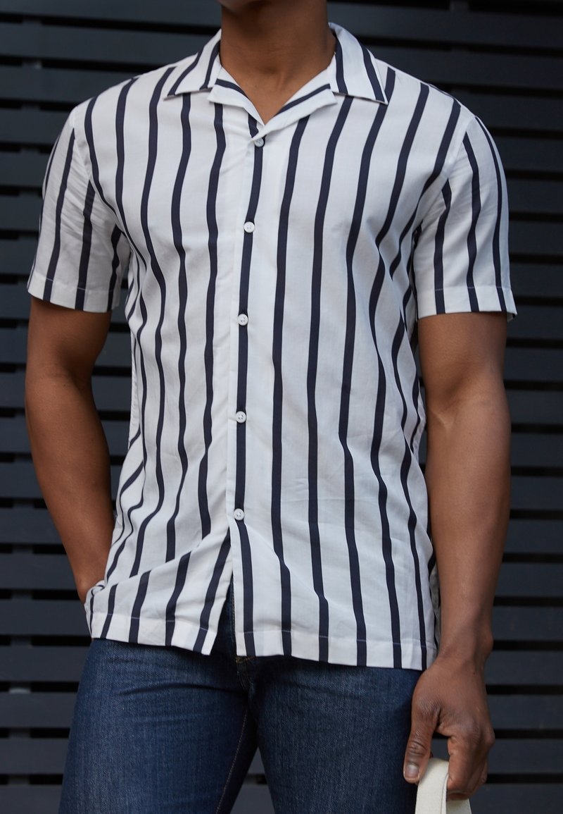 Homme portant une chemise blanche à manches courtes avec des rayures verticales noires et un jean bleu foncé, tenant un objet blanc dans sa main droite.