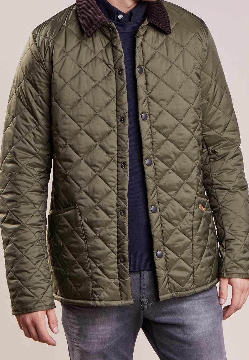 Homme portant une veste matelassée vert olive avec un col foncé par-dessus un pull bleu marine et un jean gris, debout les bras détendus.