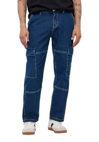 Niebieskie dżinsowe spodnie cargo charakteryzują się licznymi dużymi kieszeniami, luźnym krojem oraz kontrastowym przeszyciem. Wykończone zapięciem na guzik oraz zamek błyskawiczny.