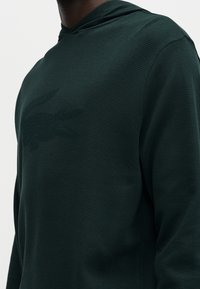 Chemise à capuche à manches longues texturée vert foncé portée par une personne devant un fond clair uni, mettant en valeur les détails du tissu et la coupe.